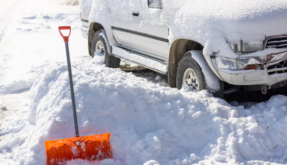 An orange snow shovel sticks out of a snowdrift in front of a snow-covered white SUV car on a sunny winter day