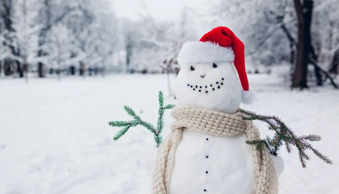 Happy snowman made outdoors in snowy winter park dressed in Santa hat, scarf with fir branches in hands. Christmas fun activities