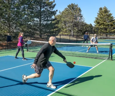 A player hits a backhand in a comptetivie doubles game of pickleball with a group of men and women on a blue and green court in spring.
