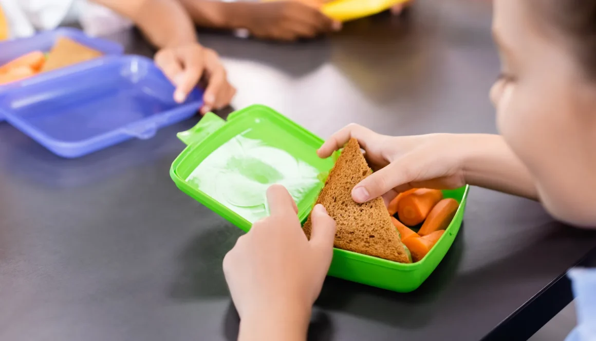 selective focus of schoolgirl taking sandwich from lunch box with fresh carrots near classmates