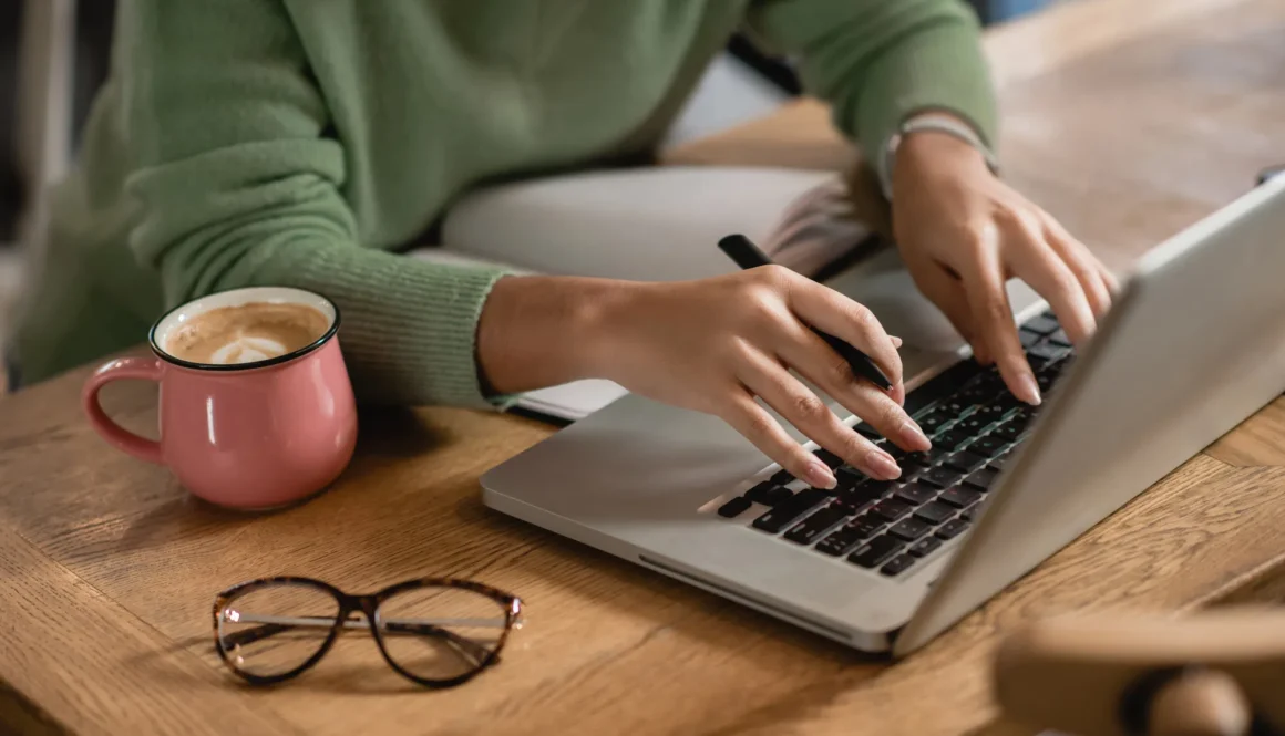 cropped view of african american woman holding pen while typing on laptop keyboard near cup of coffee